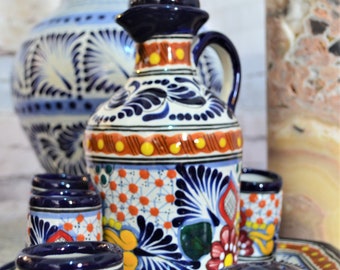 Puede incluir: Un juego de cerámica de Talavera que incluye una jarra, vasos de chupito y un salero. La cerámica está decorada con motivos florales azules, blancos, naranjas y amarillos.