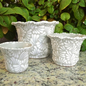 May include: Three gray ceramic flower pots with a white floral pattern. The pots are different sizes and are arranged in a row on a granite countertop.