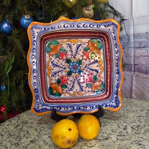 May include: A square ceramic serving dish with a blue, orange and white floral design. The dish has a scalloped edge and is decorated with intricate details.