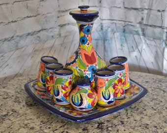 Puede incluir: Un juego de cerámica de Talavera con una jarra y seis vasos de chupito. El juego está decorado con coloridos patrones florales en tonos azules, amarillos, rojos y verdes. La jarra tiene una tapa y el juego se muestra en una bandeja cuadrada.