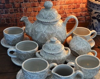 Puede incluir: Un juego de té de cerámica gris y blanco con una tetera, una jarra de crema, un azucarero y cuatro tazas y platillos. El juego presenta un delicado patrón floral.