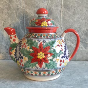 May include: A ceramic teapot with a red lid, handle, and spout. The body is decorated with a floral pattern featuring a large red poinsettia, green leaves, and colorful accents. The teapot is approximately 8 inches tall.