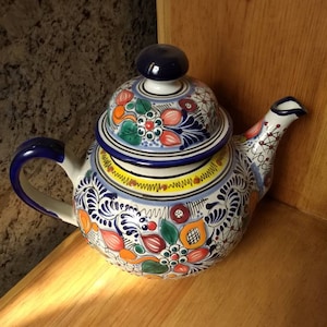 May include: A colourful ceramic teapot with a blue lid and floral designs in shades of red, yellow, green, and blue. The teapot is sitting on a wooden surface.