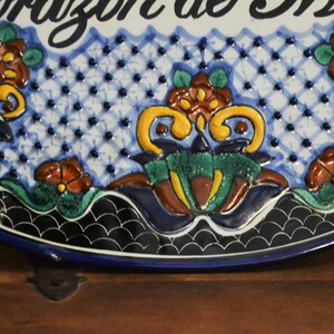 May include: A ceramic decorative plate with the words "Corazon de Mexico" in black script. The plate features a blue and white patterned background with colorful floral designs in shades of orange, green, and yellow. The plate has a dark blue border.