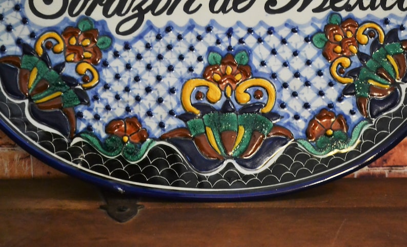 May include: A ceramic decorative plate with the words "Corazon de Mexico" in black script. The plate features a blue and white patterned background with colorful floral designs in shades of orange, green, and yellow. The plate has a dark blue border.