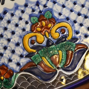 May include: Close-up of a decorative ceramic plate with a blue and white patterned background. The plate features raised floral designs in shades of yellow, green, brown, and red. The rim is a deep blue with a gold accent.