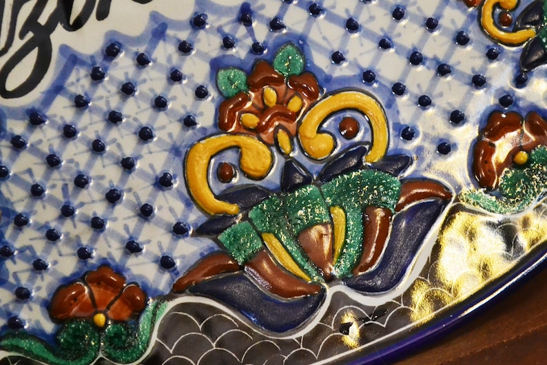 May include: Close-up of a decorative ceramic plate with a blue and white patterned background. The plate features raised floral designs in shades of yellow, green, brown, and red. The rim is a deep blue with a gold accent.