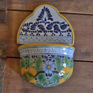 Puede incluir: Un macetero de pared de cerámica blanca con diseños florales azules y amarillos. El macetero tiene una parte superior festoneada y un fondo redondeado. Está decorado con un patrón floral en tonos azules, amarillos y verdes.