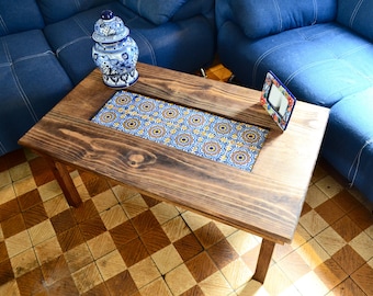 May include: A rectangular wooden coffee table with a decorative tile inlay. The table has a dark stain and a central panel of blue, yellow, and brown patterned tiles. A blue sofa is in the background, and a decorative jar and picture frame are on the table.