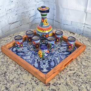 Può includere: Un set da portata in ceramica decorativa con una caraffa alta e colorata, bicchierini e un saliera. Il set è disposto su un vassoio di legno con intarsi di piastrelle floreali blu. La caraffa e i bicchierini presentano vivaci motivi floreali.