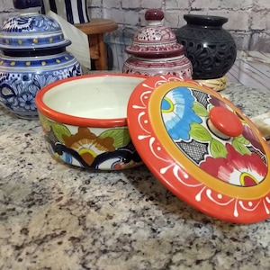 May include: A colourful ceramic bowl with a lid. The bowl is decorated with floral patterns in shades of blue, red, yellow, and green. The lid has a knob in the centre.