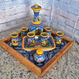 May include: A wooden serving tray with a blue and white tile design features a set of colorful ceramic shot glasses and a decanter with a matching design. The tray is perfect for serving drinks and snacks.