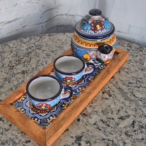 May include: A wooden tray with a colorful tile design holds three small ceramic cups and a larger ceramic teapot with a floral pattern. The cups and teapot are decorated with blue, yellow, orange, and red flowers.