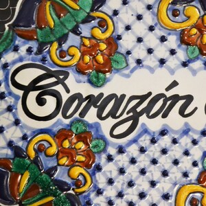 May include: Close-up of a ceramic plate with the words "Coraz&oacute;n de" in black script. The plate features a blue and white background with floral and geometric patterns in green, yellow, and brown.