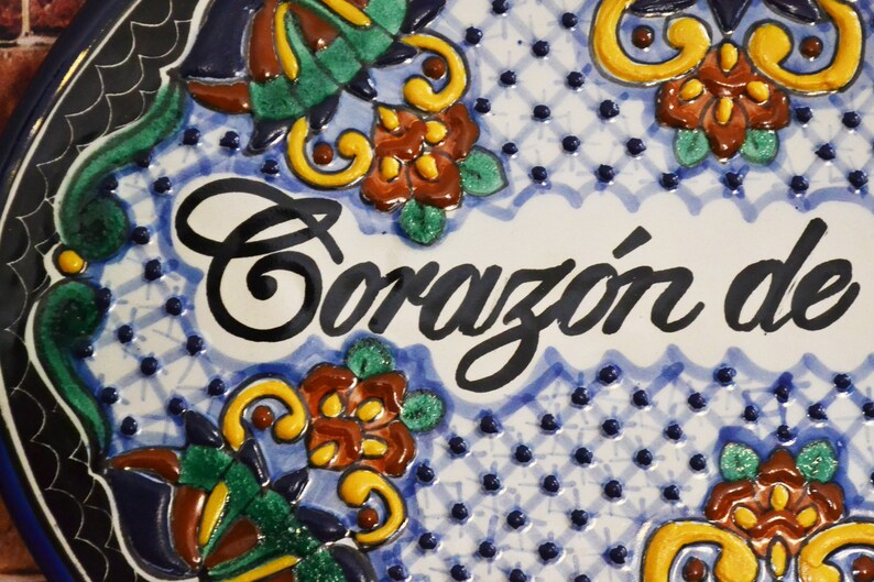 May include: Close-up of a ceramic plate with the words "Coraz&oacute;n de" in black script. The plate features a blue and white background with floral and geometric patterns in green, yellow, and brown.
