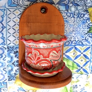 May include: A decorative wall-mounted planter with a scalloped rim. The red and pink ceramic pot features a floral design and sits on a wooden shelf. The wooden backing is stained a warm brown color, and has a hole at the top for hanging.