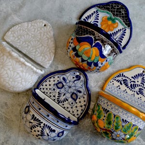 Puede incluir: Una colección de macetas de cerámica para colgar en la pared con varios diseños. Una es blanca con un estampado floral, y las otras presentan patrones florales y geométricos pintados a mano en azul, naranja y verde. Cada maceta tiene una tapa a juego.