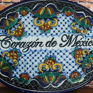 May include: Oval ceramic wall plaque with the words "Coraz&oacute;n de M&eacute;xico" in elegant script. The plaque features a blue and white background with floral and geometric patterns, and a dark blue border.