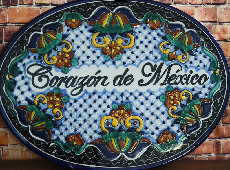 May include: Oval ceramic wall plaque with the words "Coraz&oacute;n de M&eacute;xico" in elegant script. The plaque features a blue and white background with floral and geometric patterns, and a dark blue border.