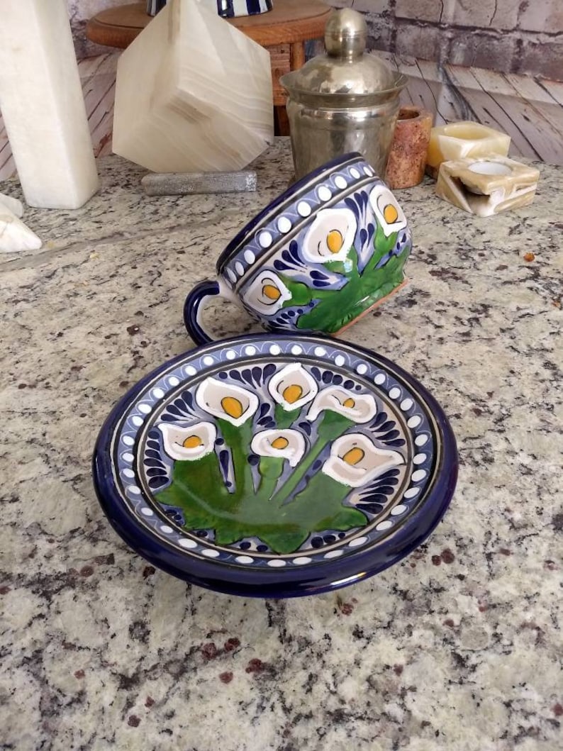 K&ouml;nnte beinhalten: Ein blaues und wei&szlig;es Keramik-Teetassen- und Untertassen-Set mit einem Blumendesign. Tasse und Untertasse sind mit wei&szlig;en Calla-Lilien mit gelben Zentren und gr&uuml;nen Bl&auml;ttern verziert. Der Hintergrund ist dunkelblau.