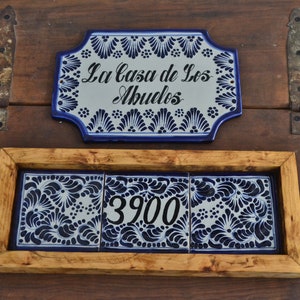 May include: A wooden frame with three blue and white ceramic tiles. The center tile has the number 3900 printed on it. Above the frame is a blue and white ceramic sign that reads "La Casa de Los Abuelos".