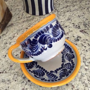 May include: A white ceramic teacup and saucer with blue and yellow floral designs. The cup has a handle and the saucer has a yellow rim.