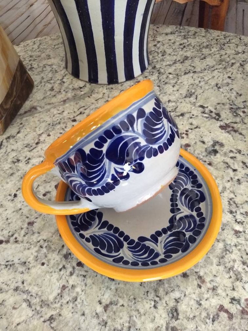 May include: A white ceramic teacup and saucer with blue and yellow floral designs. The cup has a handle and the saucer has a yellow rim.