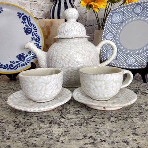 Puede incluir: Una tetera de cerámica blanca y dos tazas de té con un patrón floral blanco. La tetera tiene una tapa abovedada y un pico. Las tazas de té tienen asas. El juego está sobre una encimera de granito.