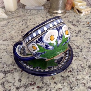 K&ouml;nnte beinhalten: Eine blaue und wei&szlig;e Keramik-Teetasse und -Untertasse mit Blumenmuster. Die Tasse hat ein gr&uuml;nes und gelbes Blumenmuster mit wei&szlig;en Bl&uuml;ten und gelben Zentren. Die Untertasse hat einen blauen Rand mit wei&szlig;en Punkten und eine wei&szlig;e Mitte mit einem blauen Blumenmuster.