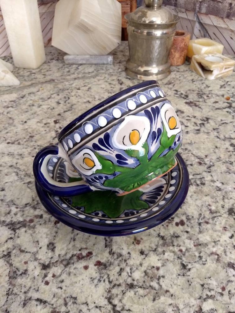 K&ouml;nnte beinhalten: Eine blaue und wei&szlig;e Keramik-Teetasse und -Untertasse mit Blumenmuster. Die Tasse hat ein gr&uuml;nes und gelbes Blumenmuster mit wei&szlig;en Bl&uuml;ten und gelben Zentren. Die Untertasse hat einen blauen Rand mit wei&szlig;en Punkten und eine wei&szlig;e Mitte mit einem blauen Blumenmuster.
