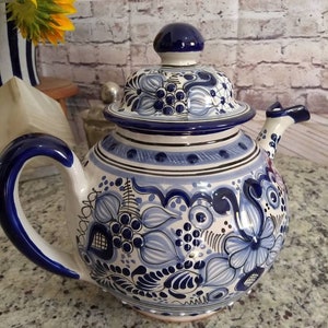 May include: A white ceramic teapot with a blue and white floral design. The teapot has a lid with a blue knob and a spout on the side. The teapot is decorated with a traditional Mexican Talavera pattern.