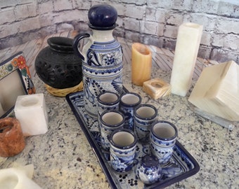 Puede incluir: Un juego de vasos de chupito de cerámica azul y blanca y una jarra a juego con tapa azul, todo en una bandeja de cerámica azul y blanca. El juego está rodeado de varios artículos decorativos, incluyendo una maceta de cerámica negra, un marco de fotos de cerámica blanca y varios candelabros blancos y beige.