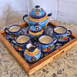 May include: A colorful Mexican Talavera pottery tea set on a wooden tray. The set includes a teapot, creamer, sugar bowl, and four teacups and saucers. The pottery is decorated with blue, yellow, and orange floral patterns.