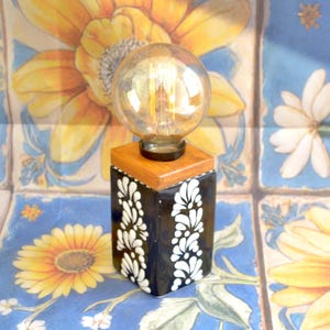 May include: A decorative table lamp with a clear globe bulb. The lamp features a wooden top and a black base adorned with white floral patterns. The lamp sits on a blue and yellow floral patterned surface.