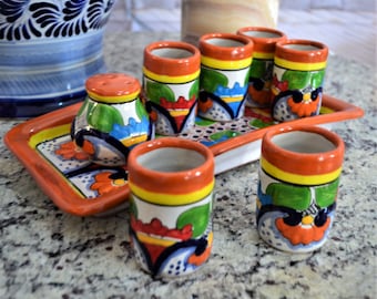 Puede incluir: Juego de seis vasos de chupito de cerámica coloridos con un diseño floral, en una bandeja a juego. Los vasos tienen un borde naranja brillante y están decorados con flores verdes, azules, amarillas y rojas.