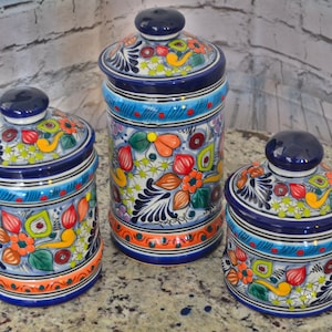 Puede incluir: Conjunto de tres botes de cerámica coloridos con tapas azules y diseños florales. Los botes están decorados con colores brillantes, incluyendo naranja, amarillo, verde y azul.