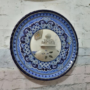 May include: A blue and white ceramic mirror with a decorative floral design. The mirror is round and has a scalloped edge. The mirror is reflecting a white ceramic jar with a decorative design.