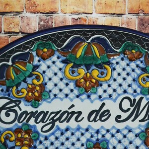 May include: A ceramic plate with a blue border and the words "Coraz&oacute;n de M&eacute;xico" in elegant script. The plate features a white and blue patterned background with colorful floral designs in yellow, brown, and green. The background is a brick wall.
