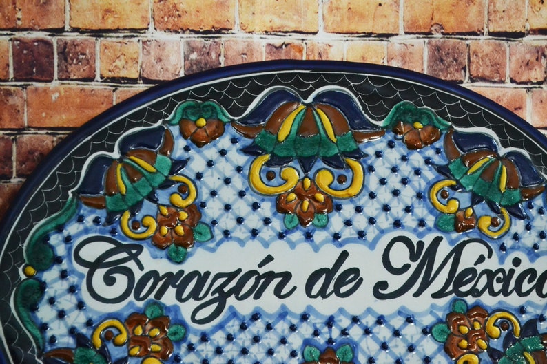 May include: A ceramic plate with a blue border and the words "Coraz&oacute;n de M&eacute;xico" in elegant script. The plate features a white and blue patterned background with colorful floral designs in yellow, brown, and green. The background is a brick wall.