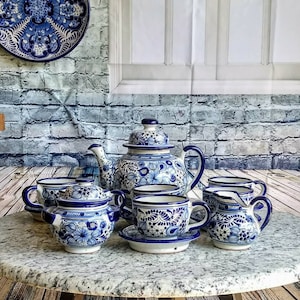 Puede incluir: Un juego de té de cerámica Talavera azul y blanco, con tetera, jarra de crema, azucarero y dos tazas y platillos. El juego presenta intrincados diseños florales y detalles azules.