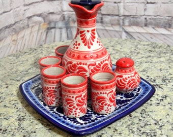 Puede incluir: Un juego de seis vasos de chupito de cerámica pintados a mano y una jarra a juego con un diseño floral azul y rojo. El juego se muestra en una bandeja de cerámica azul y blanca.