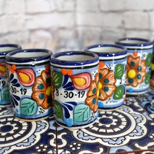 May include: Six colourful ceramic shot glasses with floral designs and the date "3-30-19" painted on them. The glasses are arranged in a row on a tiled surface.