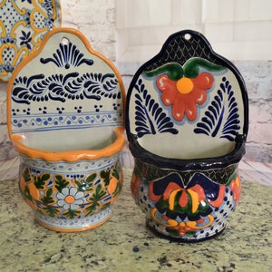 Puede incluir: Dos macetas de pared de Talavera con diseños florales coloridos. La maceta de la izquierda tiene un diseño azul y blanco con acentos naranja. La maceta de la derecha tiene un diseño azul y negro con acentos naranja, rojo y verde.