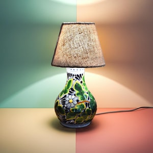 May include: A decorative table lamp with a ceramic base featuring a floral design in green, blue, and white. The lamp has a beige fabric shade and a power cord. The lamp is a decorative home decor item.