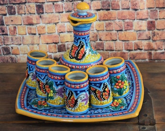 May include: A colorful ceramic tequila set with a pitcher and six shot glasses. The set features a blue background with yellow, orange, and green accents. The pitcher and glasses are decorated with butterfly designs.