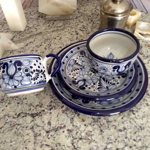 May include: A set of blue and white Talavera pottery includes a cup and saucer with a floral design. The cup and saucer are on a larger plate with a similar design.
