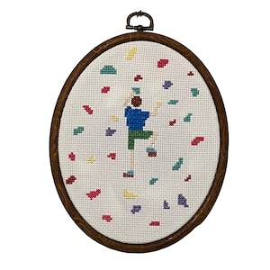 Op de afbeelding: Ovale kruissteekborduurwerk met een figuur die klimt met kleurrijke confetti-achtige vormen. De figuur draagt een blauw shirt en een groene broek. De borduursel zit in een houten hoepel met een metalen sluiting.