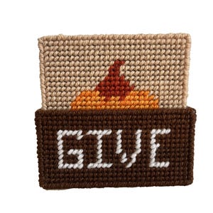 May include: A brown and beige cross-stitch with the word "GIVE" in white letters. The top part of the cross-stitch features a stylized orange and red flame.