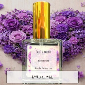 May include: A perfume bottle with a gold cap and a label that reads "Cake & Barrel Spellbound Eau De Parfum 1 oz." is centered in front of a heart-shaped arrangement of purple flowers. The text "L*VE SP*LL" is at the bottom.