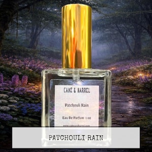 May include: A clear glass perfume bottle with a gold cap. The label reads "Cake & Barrel Patchouli Rain Eau De Parfum 1 oz." The background is a blurred image of a forest path with purple flowers.